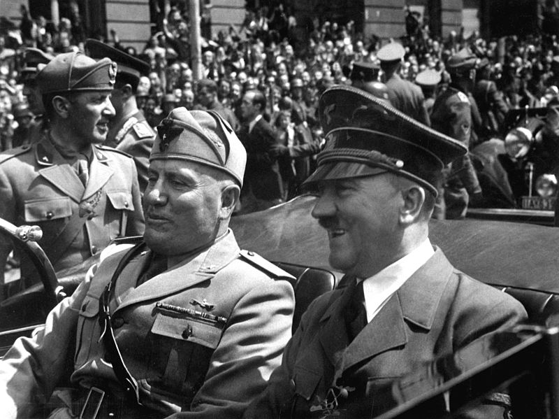 To smilende menn med militæruniformer kjører sammen i en åpen bil. Soldater og en stor folkemengde i bakgrunnen. Svart-hvitt foto.  