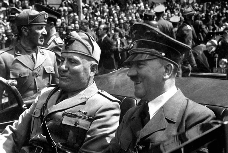 To smilende menn med militæruniformer kjører sammen i en åpen bil. Soldater og en stor folkemengde i bakgrunnen. Svart-hvitt foto.