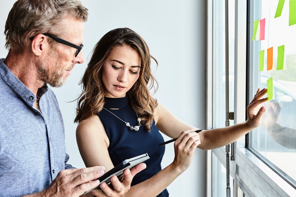 To personer samarbeider om å skrive noe på ulike post-it-lapper. Den ene personen henger opp post-it-lapper på en vindusflate. Foto.