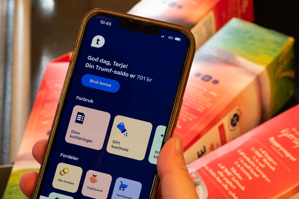 Ei hånd holder en mobiltelefon. Skjermen på mobilen viser framsida til fordelsprogrammet Trumf sin medlemsapp. Foto.