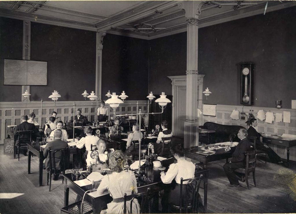Kvinner og menn sitter på rekke ved flere bord. Til høyre sitter det en mann som ser ut til å overvåke det hele. Personene på bildet er telegrafister ved Bergen telegrafstasjon i 1899. Foto.