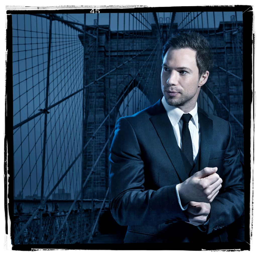 Mann i dress poserer med Brooklyn bridge i bakgrunnen. Brukonstruksjonen danner tydelige linjer og mønstre. Foto.