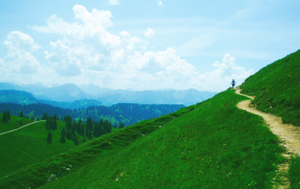 Sti i grønt alpelandskap. En person er på fjelltur. Nydelig utsikt mot en blå horisont. Foto.