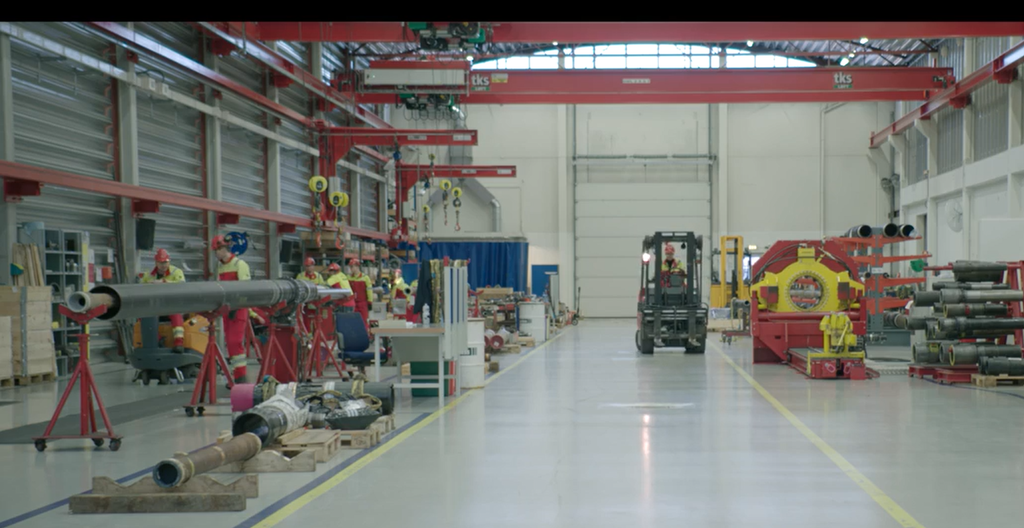 Utstyr som skal sendes offshore, klargjøres i et verksted i en stor hall. Flere rør og arbeidere, en av arbeiderne kjører gaffeltruck. Foto.