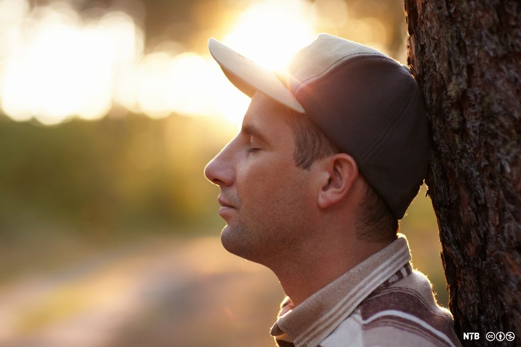 Ein ung mann med attlatne auge støttar hovudet inntil stammen på eit tre. Foto.