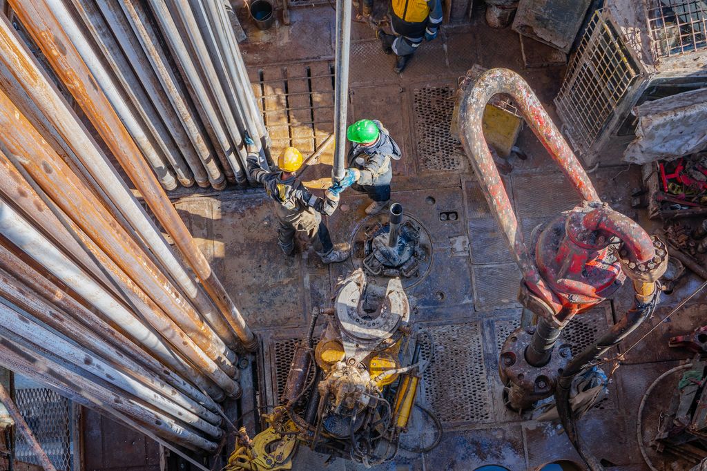 Boredekk sett ovenfra. Til venstre vises rør som lagres stående på boredekk. I midten er to arbeidere i ferd med å håndtere en rørlengde som henger fra toppen av boretårnet. Foto.