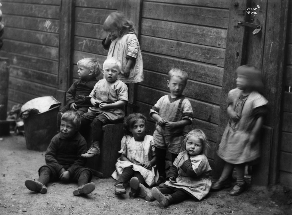 Åtte småunger foran en uthusvegg i en bakgård. Miljøet virker fattigslig. 1920-tallet. Foto.