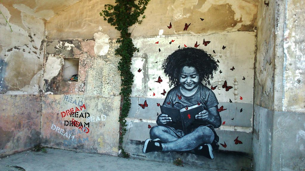 Graffiti på en mur av ei jente som sitter på bakken med beina i kors og leser i ei bok. Hun smiler. Mange røde sommerfugler svermer rundt henne. Ved siden av jenta står det "READ" og "DREAM" flere ganger om hverandre. Foto.