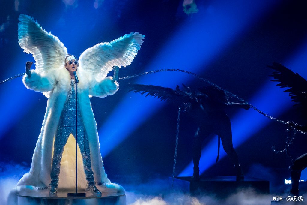 Mann står foran en mikrofon på en scene og synger. Han har på seg glitrende overdel og bukse, solbriller og en hvit pelskappe som når ned til gulvet. Han har store englevinger, og begge håndleddende hans er lenket til svartkledde dansere med svarte vinger og horn. Foto.