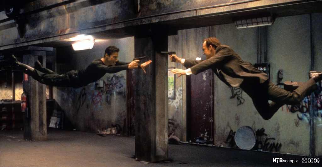 To menn svever liggende i løse luften. De peker mot hverandre med en pistol. Foto.