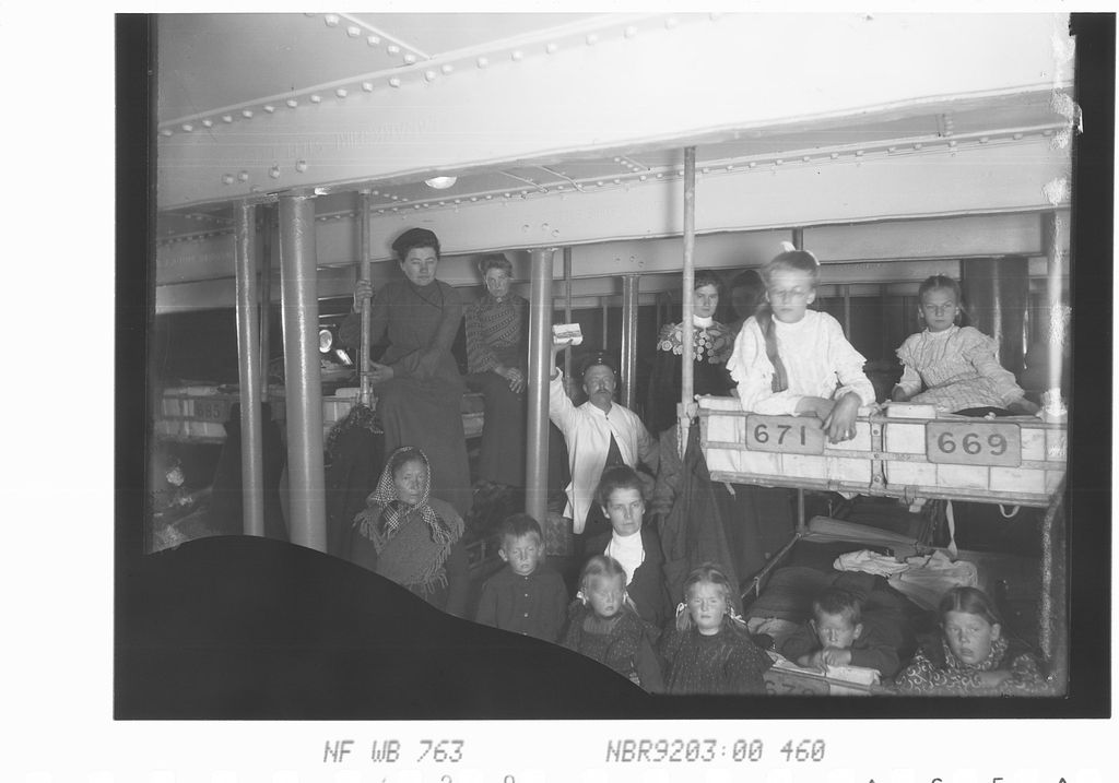 Mange voksne og barn stuet sammen i et skipsrom. To av barna sitter i køyer. Foto. 