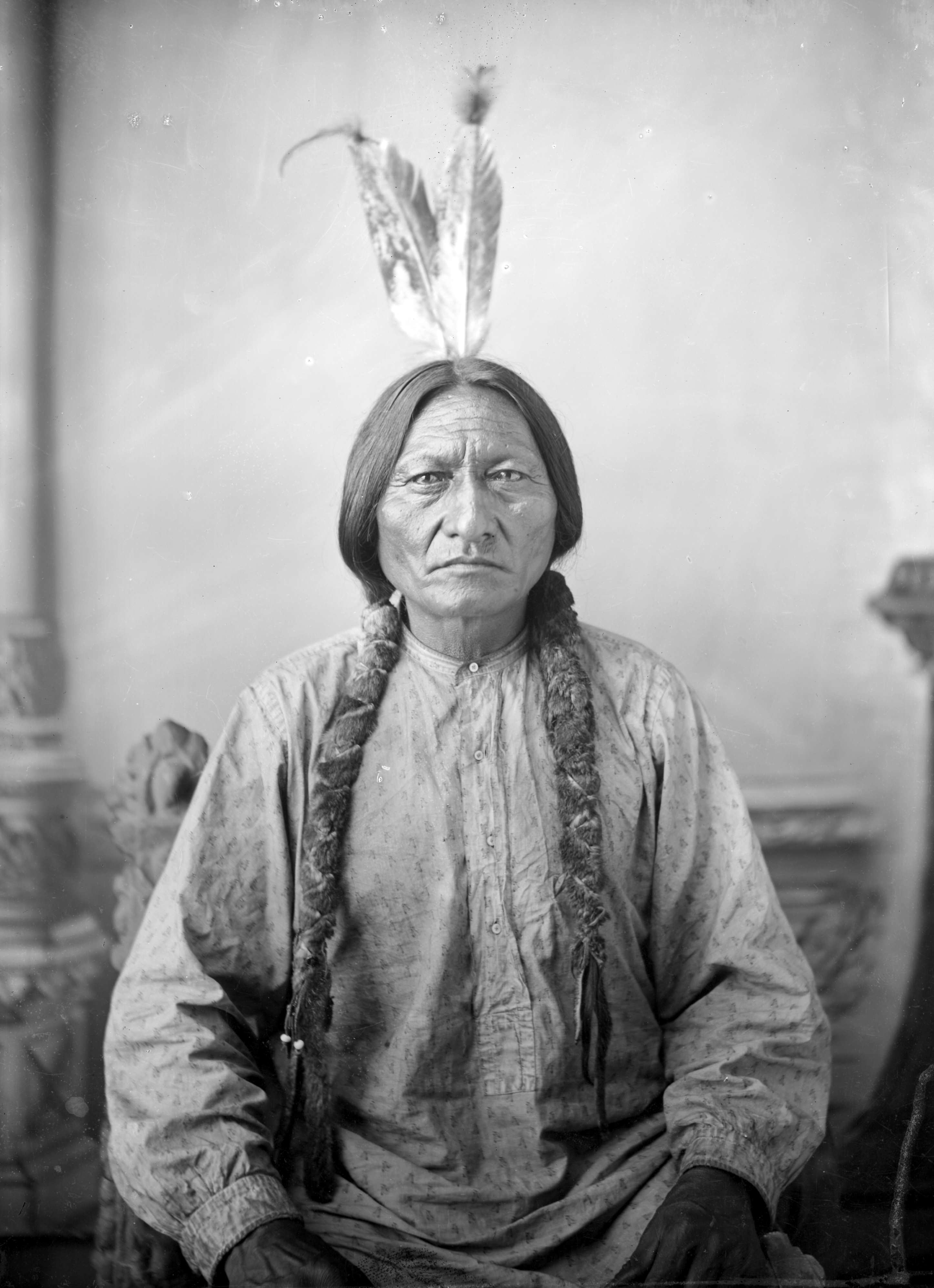 Photo: Portrait of Sitting Bull. We see an older, Native American man. His hair is in two thick braids. He wears a patterned shirt and has two feathers in his hair. 