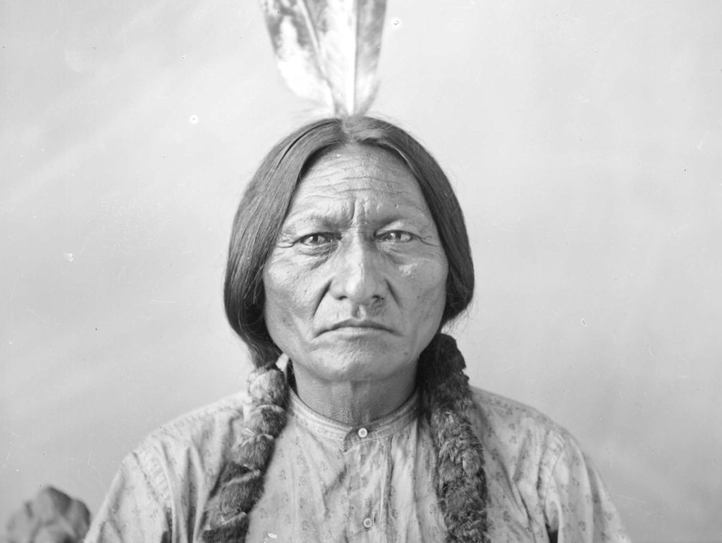 Photo: Portrait of Sitting Bull. We see an older, Native American man. His hair is plaited in two thick braids. He is wearing a patterned shirt, and has two feathers in his hair.