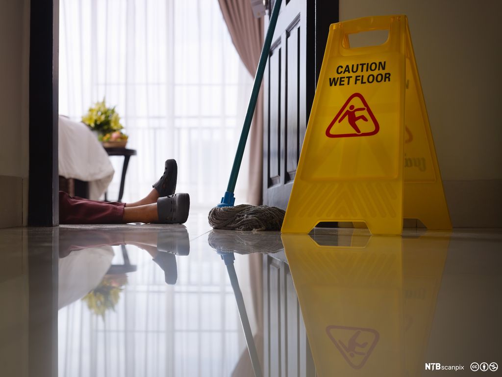 Det vises føttene og litt av beina til en person som ligger på gulvet. Ved siden av står det en vaskekost og et gult skilt med påskriften "CAUTION WET FLOOR". Foto.