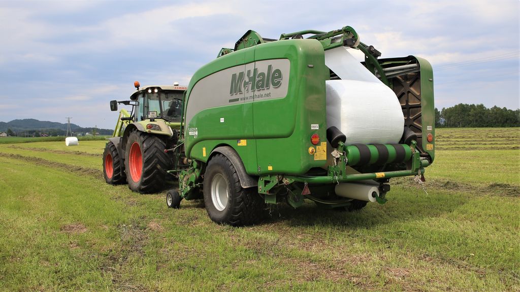 Traktor med ei kombipresse på et jorde. Bak i pressa blir en rundballe pakket inn i kvit, bred plast. Foto.