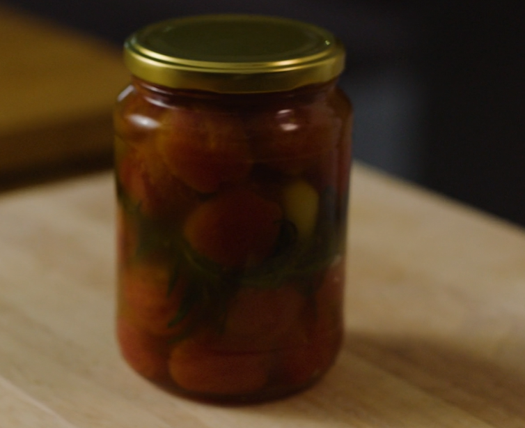 Et glass med bakte småtomater i olje og kvister av friske krydderurter. Foto.