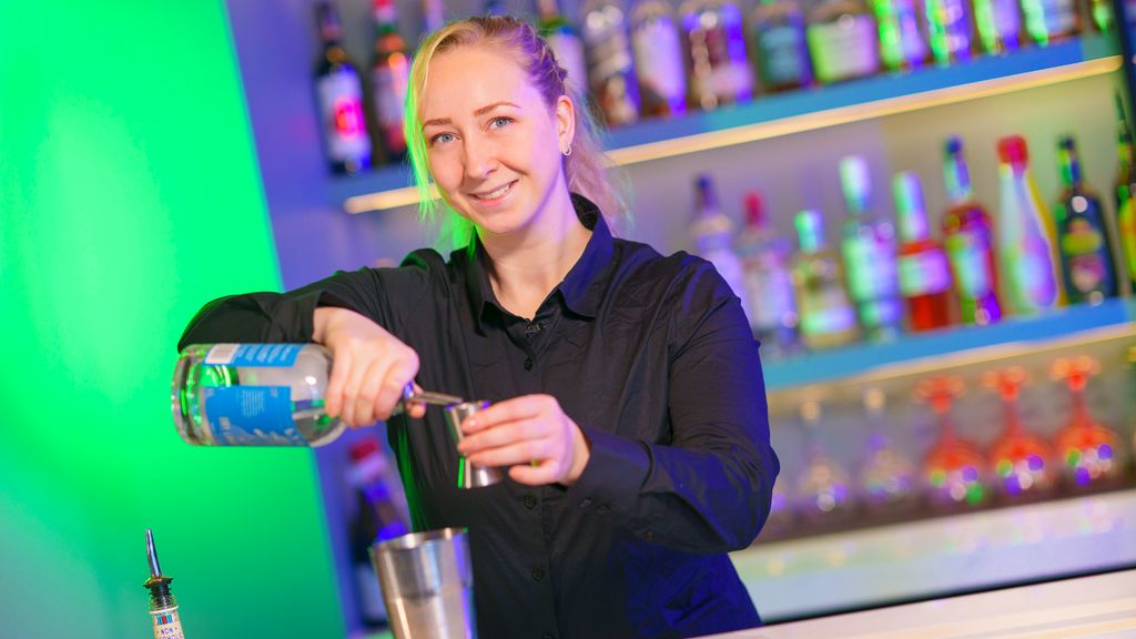 Smilende bartender bruker målebeger. Rekker med flasker i opplyste hyller i bakgrunnen. Foto.
