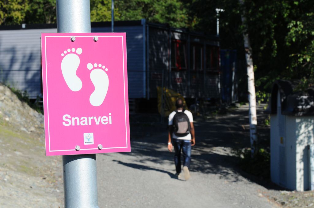 Eit skilt med illustrasjon av to føter og teksten "snarvei", framfor ein grussti. I bakgrunnen er det ein mann som går på grusstien. Foto. 