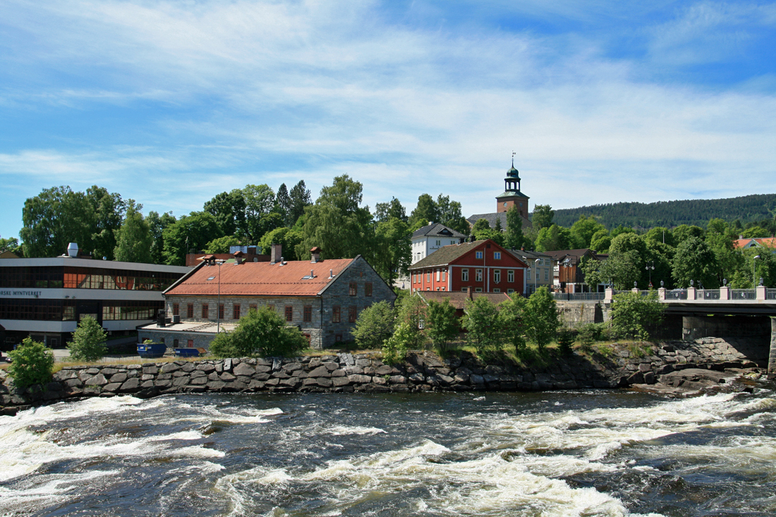 Moderne bygning, nokre bygningar frå 1800-talet og ei kyrkje i bakgrunnen. Elv med stryk i forgrunnen. Foto.