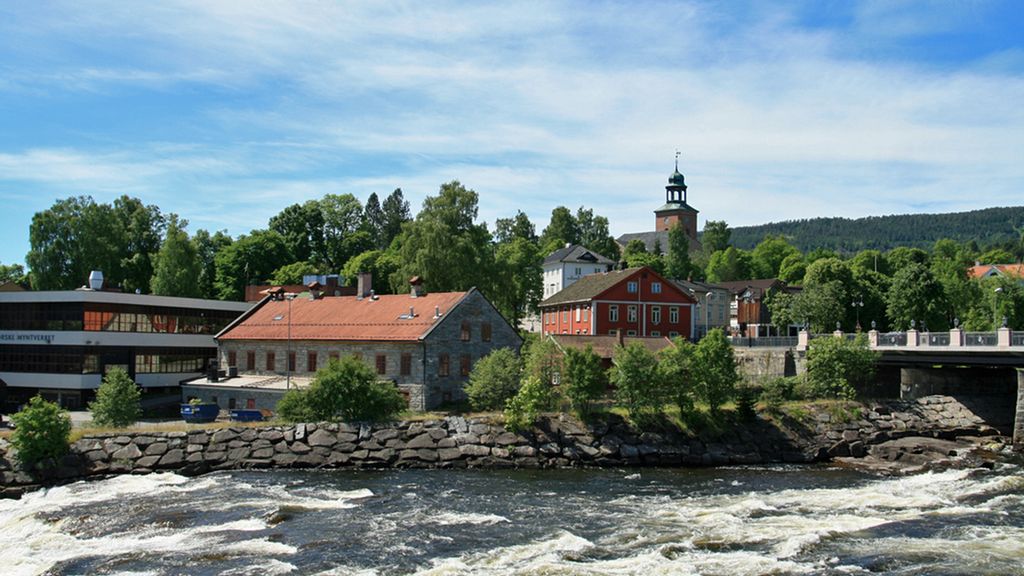 Moderne bygning, nokre bygningar frå 1800-talet og ei kyrkje i bakgrunnen. Elv med stryk i forgrunnen. Foto.