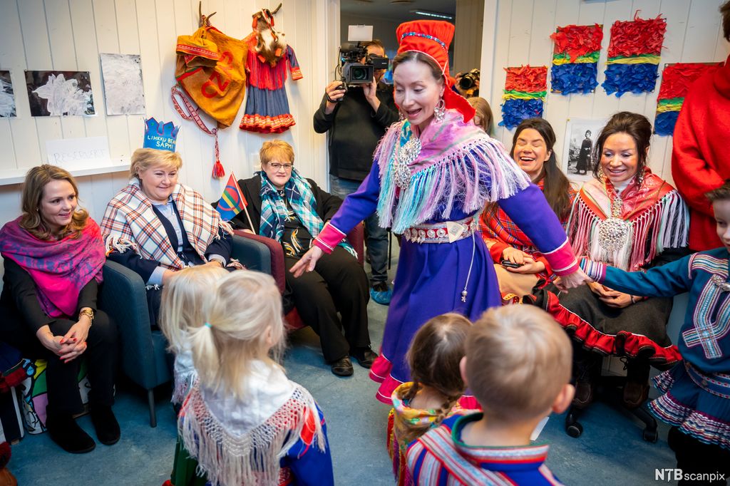 En kvinne og barn kledd i tradisjonelle samiske drakter er med på en forestilling. Linda Hofstad Helleland, Erna Solberg, Trine Skei Grande og to kvinner i samiske drakter sitter og ser på. En kameramann står i bakgrunnen. Foto. 