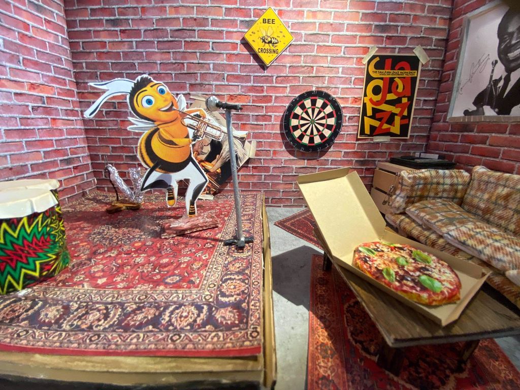 Ein teikneseriefigur som ser ut som ein glad drone (hannbie) med menneskelege trekk, speler trompet i eit rom som er innreidd med scene og sofa med bord. Foto av ein modell.
