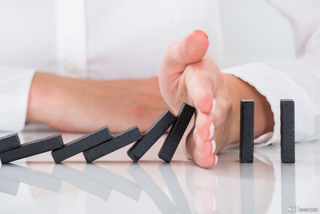 Dominobrikker på rekke. De seks første brikkene har falt. Bak den sjette brikken kommer en hånd inn og sperrer, slik at resten av brikkene i rekka blir stående. Foto.