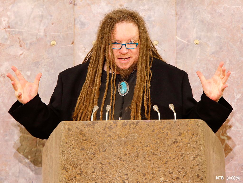 Jaron Lanier, forfatter, musiker og forsker, på en talerstol med flere mikrofoner foran seg. Han er svartkledd, med langt hår og briller. Foto.