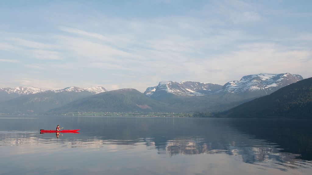 Ein raud kajakk på ein fjord med fjell i bakgrunnen. Det er litt snø på fjella. Foto.