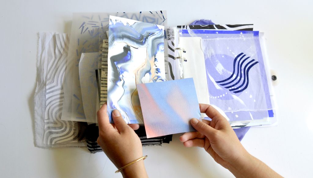 Hender som blar i en bok med ulike tekstil og fargeprøver. Foto. 