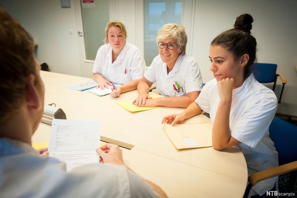 Personer med hvit arbeidsuniform med papirer foran seg sitter rundt et bord. Foto.