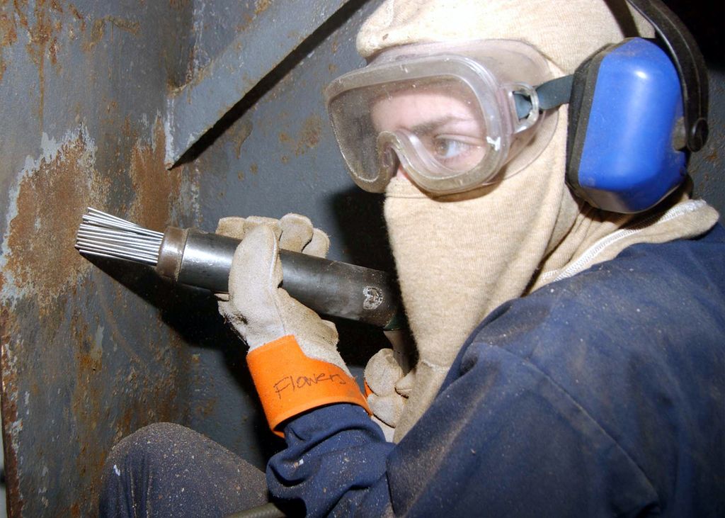 Ein person med arbeidstøy og verneutstyr held ein trykkluftdriven nålpistol mot ei metalloverflate som har fått korrosjon. Foto.