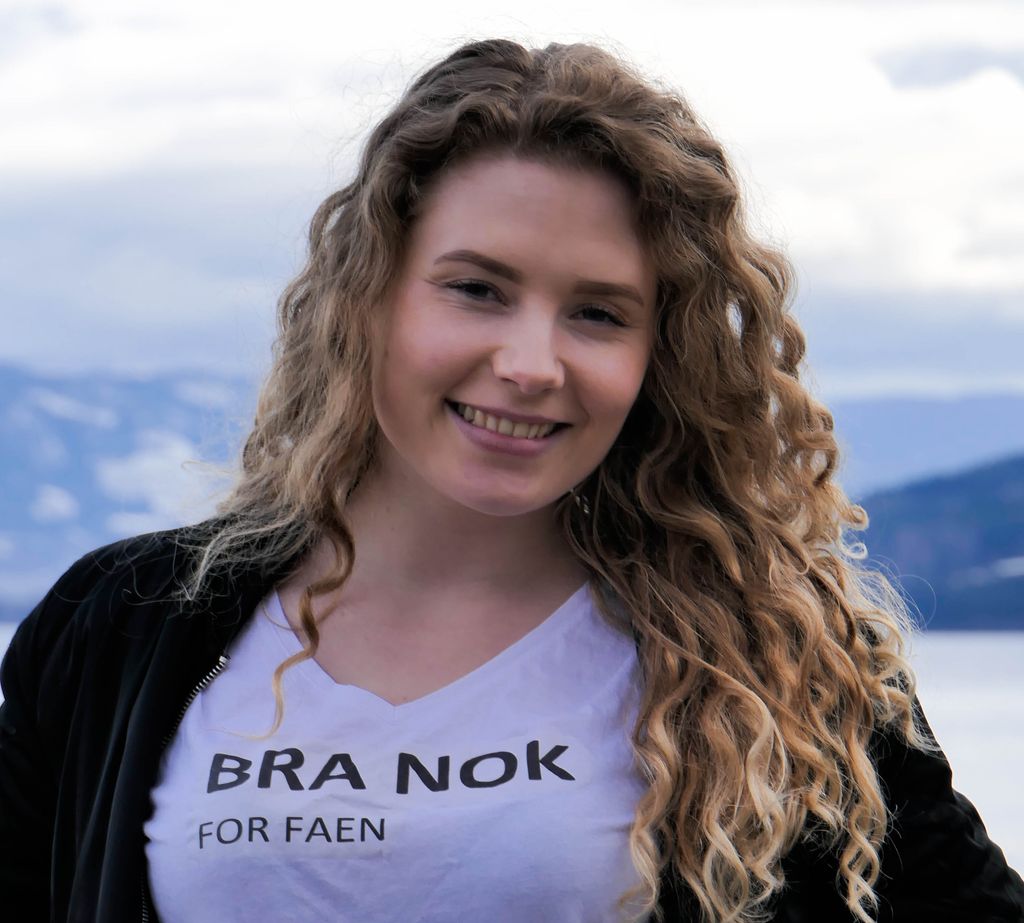 Ei ung kvinne poserer for kamera med ei T-skjorte der det står "Bra nok – for faen". Foto.