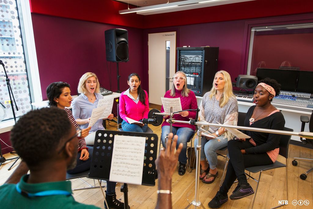 Photo: A choir of women are rehearsing. A man is conducting. 