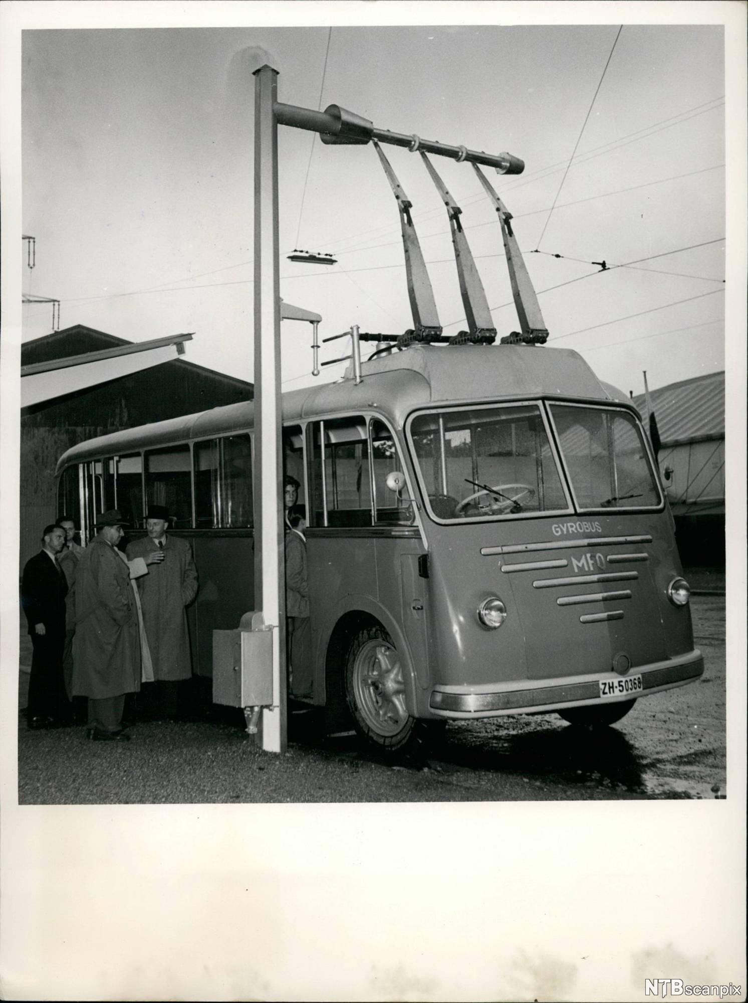 Svarthvittbilde fra 1950-tallet av menn i frakk ved en elektrisk buss. Foto.