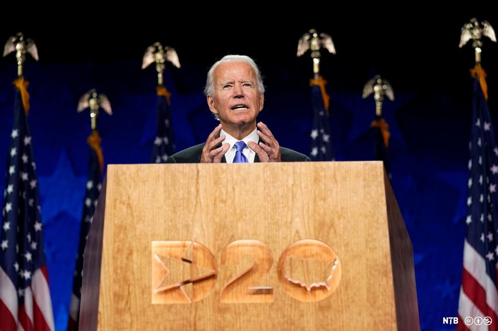 Joe Biden standing behind a wood pulpit with 'D20' engraved on it. Behind Biden, there are six American flags, each with a metal eagle on top. Joe Biden is an elderly, white-haired man, wearing a suit and a blue tie. Photo.