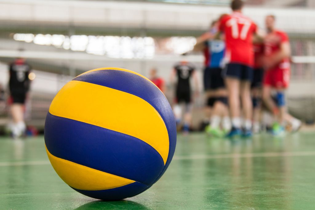 Gul og blå volleyball som ligger i ro på grønt gulv. Volleyballspillere og volleyballnett vises diffust i bakgrunnen. Foto.