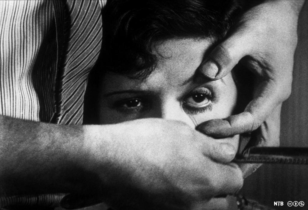 Nærbilde av hodet til en kvinne. En mann holder henne bakfrag og sperrer opp et av øynene hennes med den ene hånda. I den andre hånda holder han opp en barberkniv. Svart-hvitt-foto.