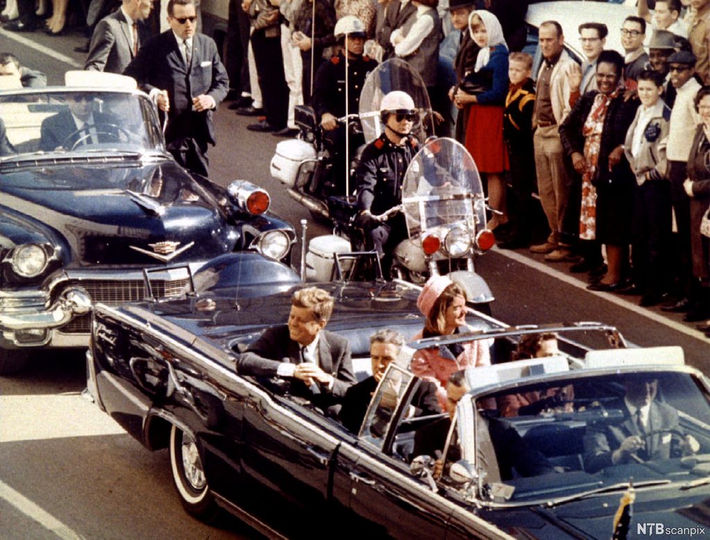 En smilende president John F. Kennedy og kona Jackie sitter i baksetet på en åpen bil som kjører i en kortesje med sikkerhetsvakter rundt. Langs veikanten står folk tett i tett og ser på. Foto.