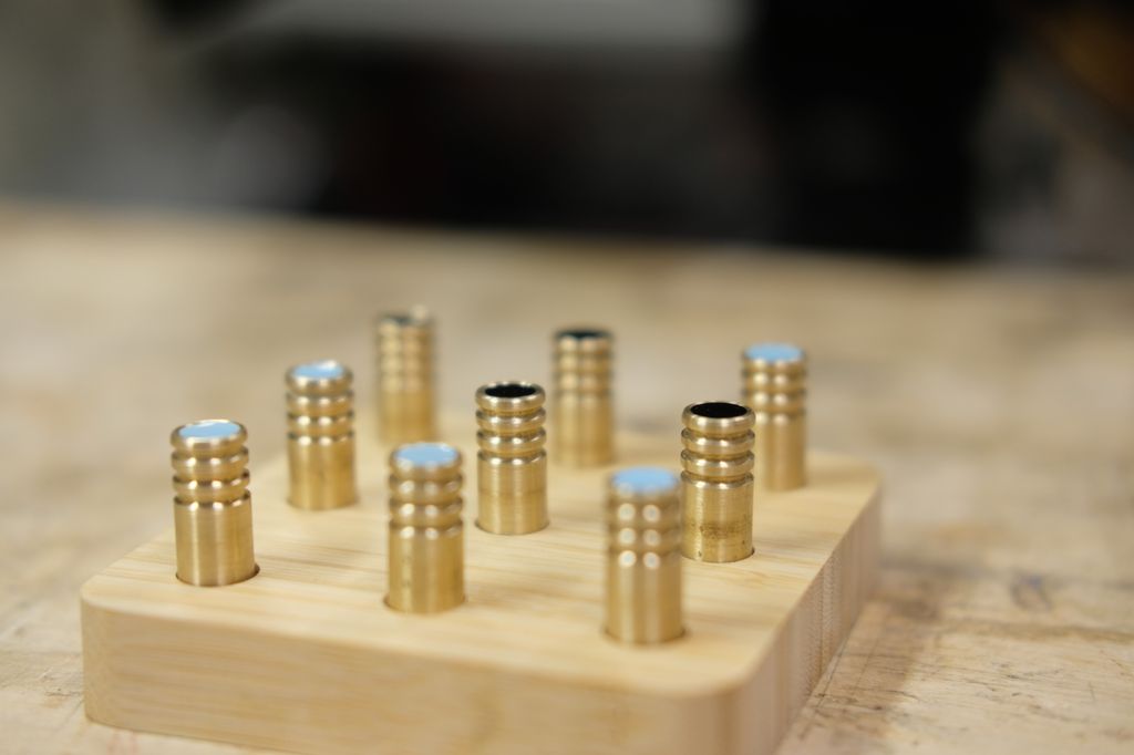 Tre-på-rad-spillebrett i bambus med dreide brikker i messing. Brikkene har dreide spor og er merka med lyseblått eller svart i en forsenking på toppen. Foto.