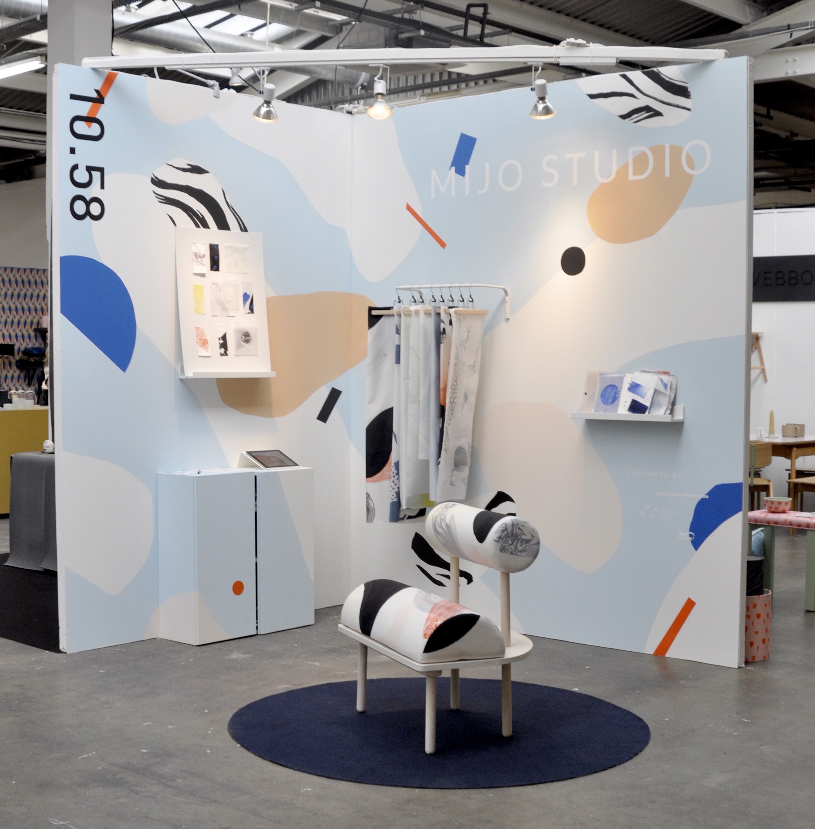 Møbler og tekstiler i ulike farger stilt ut på en messestand. Det er en kreativt utformet stol, et gulvteppe, et skap, ei tavle, ei vegghylle og stoffprøver som henger på et stativ som er festet til veggen. Foto.