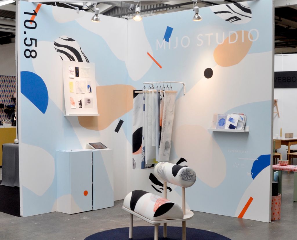 Møblar og tekstilar i ulike farger stilte ut på ein messestand. Det er ein kreativt utforma stol, eit golvteppe, eit skap, ei tavle, ei vegghylle og stoffprøver som heng på eit stativ som er festa til veggen. Foto.