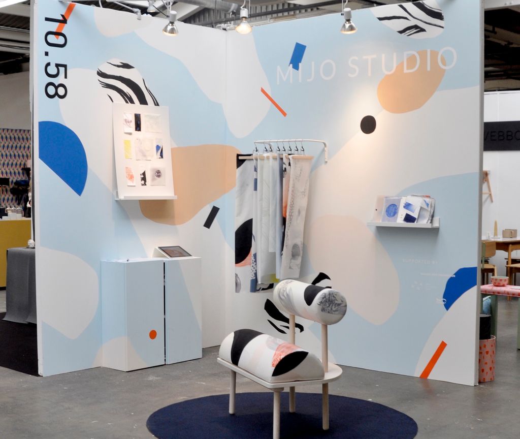 Møbler og tekstiler i ulike farger stilt ut på en messestand. Det er en kreativt utformet stol, et gulvteppe, et skap, ei tavle, ei vegghylle og stoffprøver som henger på et stativ som er festet til veggen. Foto.