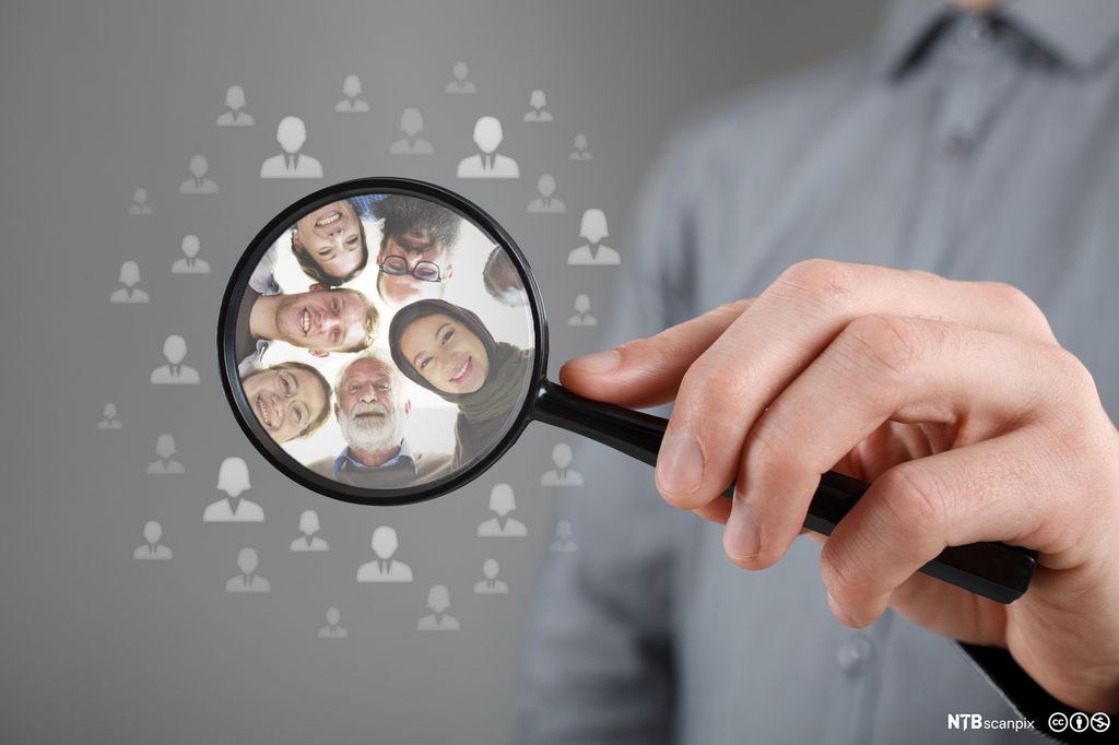 Hånd som holder forstørrelsesglass med  flere mennesker i. Foto.