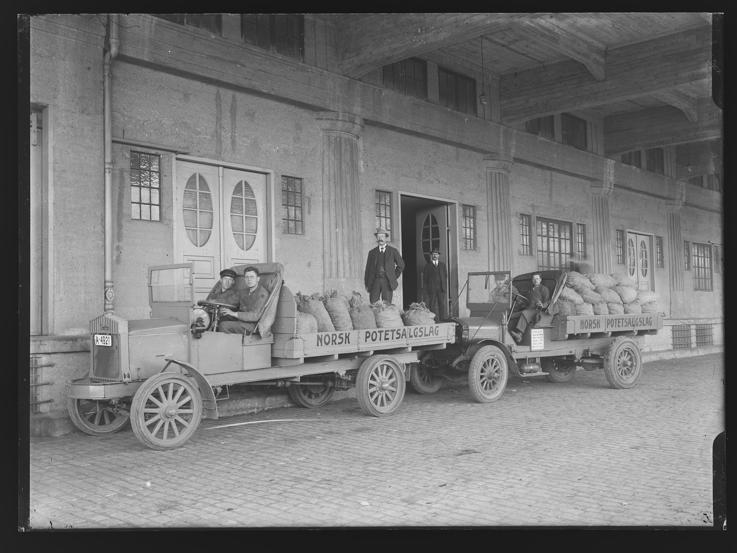 Historisk svart-hvitt bilde av biler med påskriften Norsk potetsalgslag og potetsekker på lasteplanene. Foto.