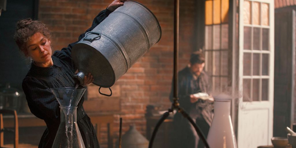 En kvinne i kjole heller væske fra en stor metallbeholder over i en glasskolbe. Ved siden av henne står en annen glasskolbe der hvit damp kommer ut. I bakgrunnen står en mann og leser. Foto.