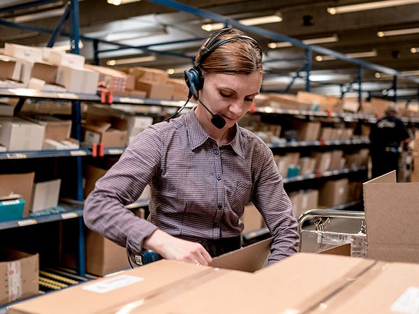 Dame med hodesett pakker varer på et lager. Foto.