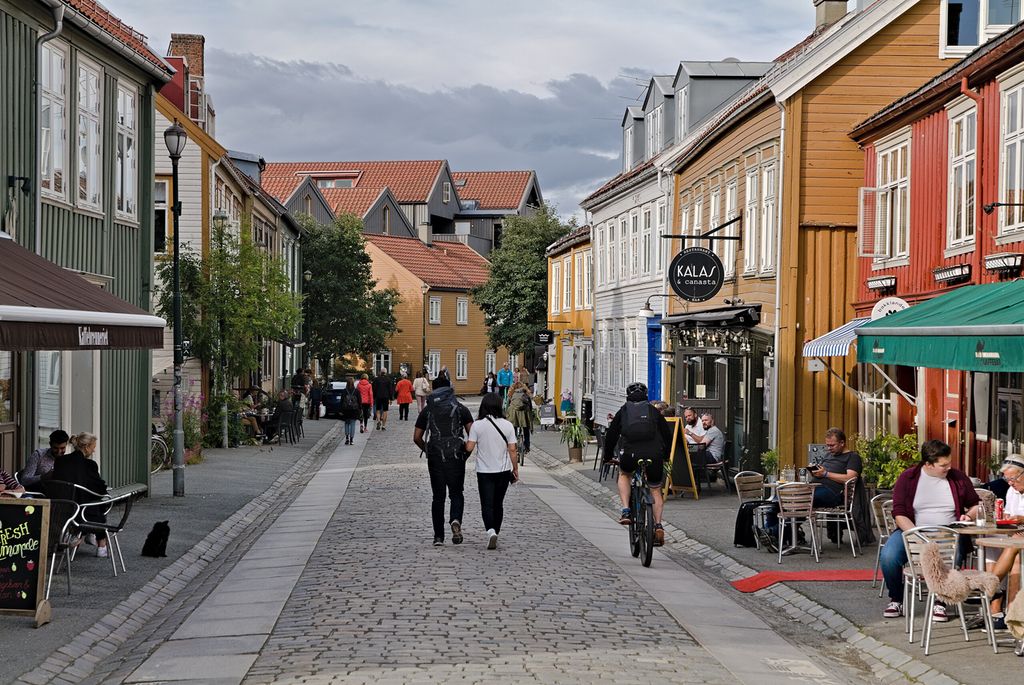 Ei gate med brostein og godt vedlikeholdte gamle hus på hver side. Noen personer sitter på fortauskafeer. Noen går i gata, mens andre sykler. Foto. 