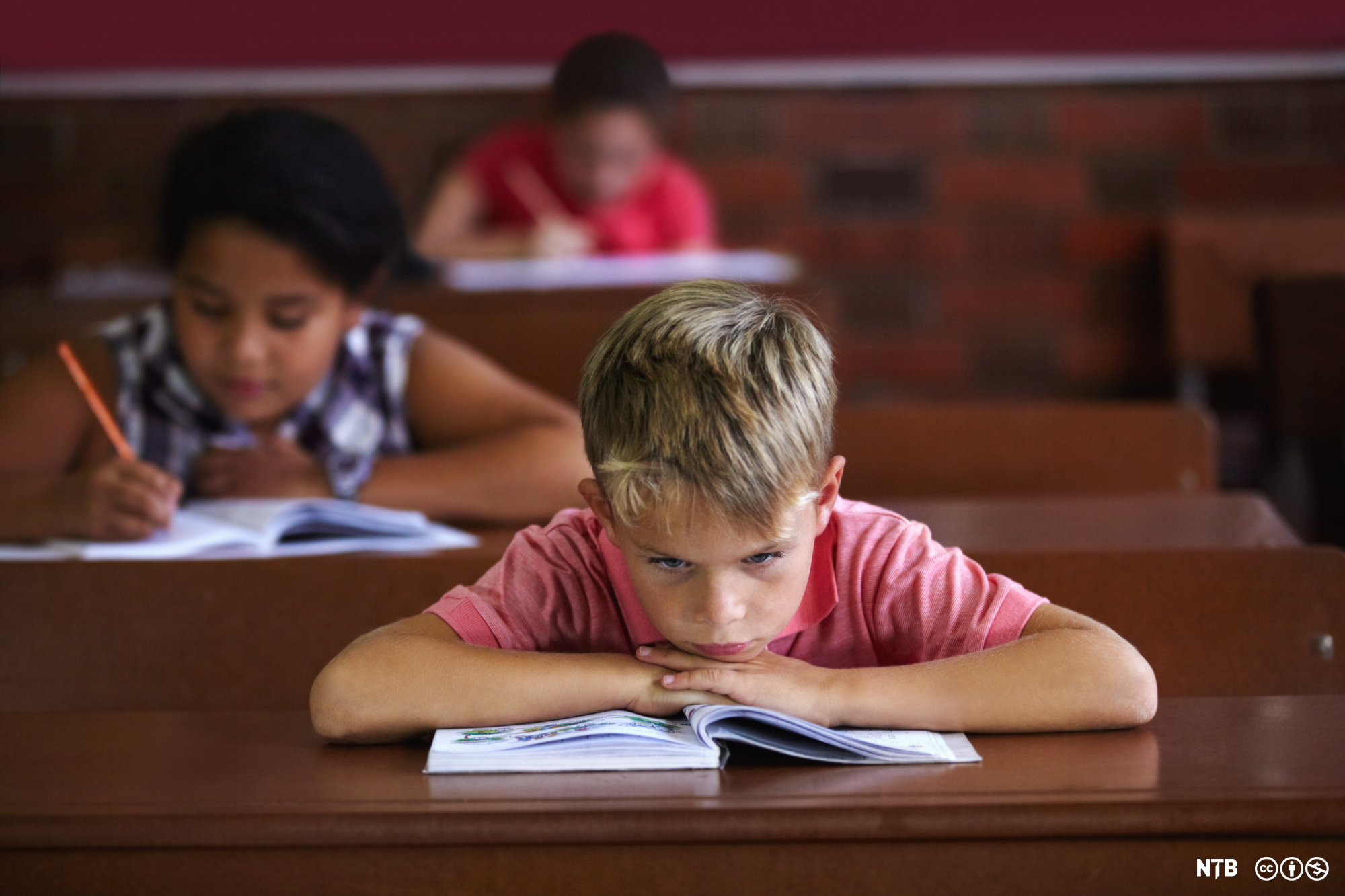 A young boy is sitting by a school desk, looking frustrated and bored. There are two other students in the background, working. Photo.