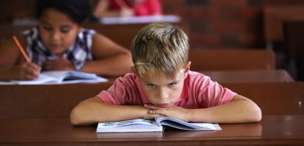 A young boy is sitting by a school desk, looking frustrated and bored. There are two other students in the background, working. Photo.